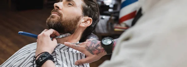 Tätowierter Barbier rasiert Bart eines brünetten Mannes mit Rasiermesser im Friseursalon, Banner — Stockfoto