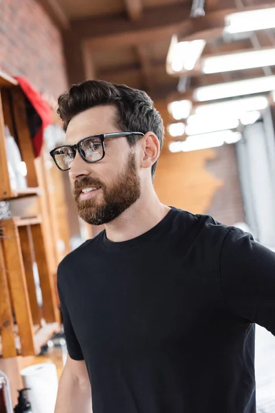 Bearded barber in eyeglasses and black t-shirt standing in barbershop — Stock Photo