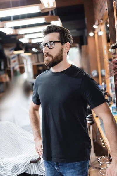 Peluquería en gafas mirando hacia otro lado cerca de sillón y la capa de peluquería en la barbería - foto de stock