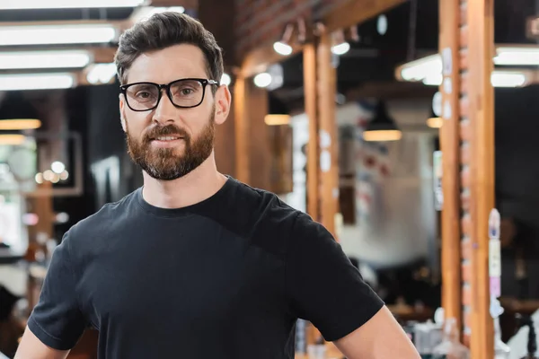 Lächelnder Friseur mit Brille blickt in verschwommenem Schönheitssalon in die Kamera — Stockfoto