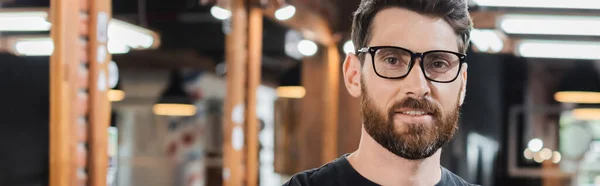 Portrait of brunette hairdresser in eyeglasses looking at camera in blurred barbershop, banner — Stock Photo