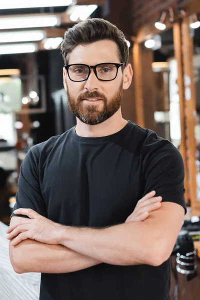 Peluquero barbudo en gafas cruzando brazos y mirando la cámara en la barbería - foto de stock