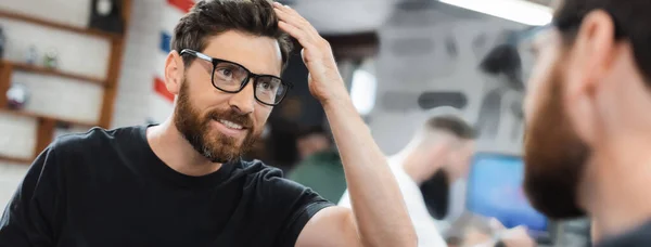 Cheerful client in eyeglasses looking at hair near mirror in barbershop, banner — Stock Photo