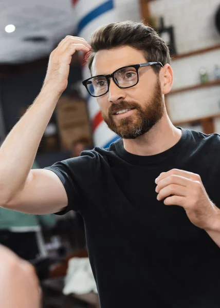 Uomo sorridente in occhiali che tocca capelli vicino a specchio in negozio di barbiere — Foto stock