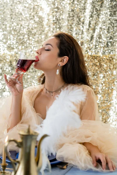 Femme glamour en robe de tulle boire du vin rouge près flou cruche de cuivre sur fond de paillettes — Photo de stock
