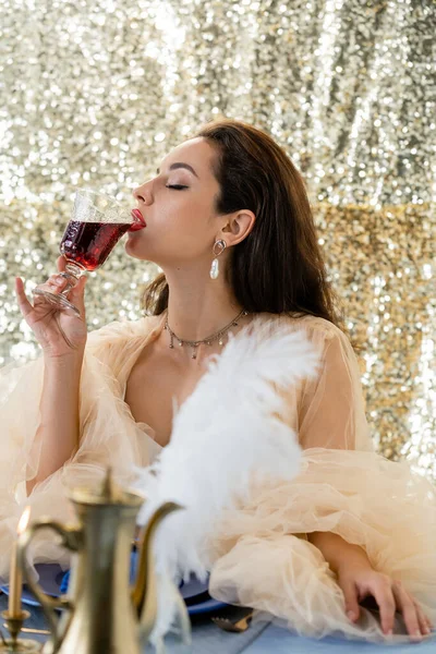 Mujer glamour en vestido de tul beber vino tinto cerca borrosa jarra de cobre sobre fondo brillo - foto de stock