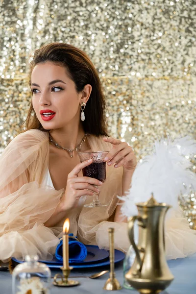 Verführerische Frau im Chiffonkleid mit Rotwein in der Hand und Blick weg neben brennender Kerze auf Hintergrund mit Glitzer — Stockfoto