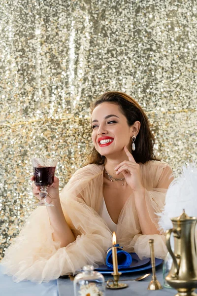Femme heureuse dans une élégante robe en tulle tenant un verre de vin rouge et regardant loin sur fond brillant — Photo de stock