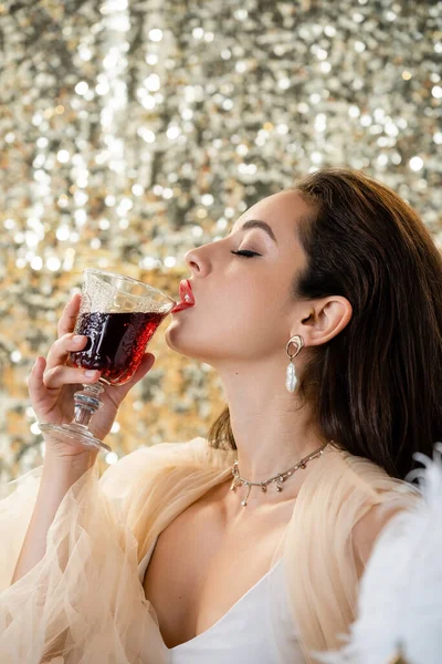 Соблазнительная женщина в шифонском платье пьет красное вино на блестящем серебряном фоне — стоковое фото