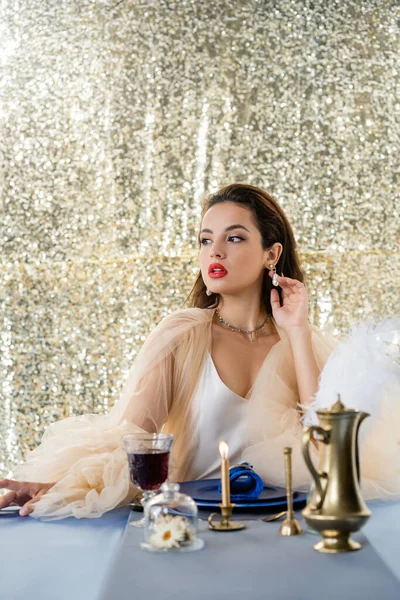 Morena mujer en vestido elegante mirando lejos cerca de vino tinto y vela ardiente sobre fondo brillante - foto de stock