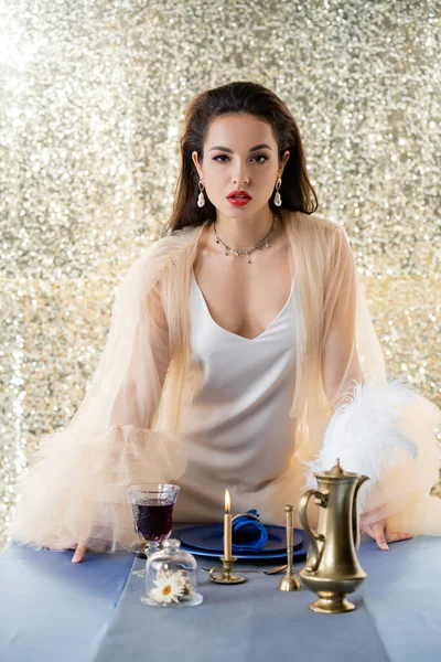 Mujer joven en vestido elegante mirando a la cámara cerca de platos azules y una copa de vino sobre fondo plateado brillo - foto de stock