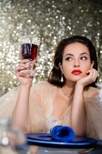 Brunetta elegante donna in tulle vestito in possesso di un bicchiere di vino rosso vicino piatti blu su sfondo glitter — Foto stock