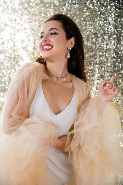 Mujer morena feliz en vestido elegante y maquillaje mirando hacia otro lado sobre fondo plateado brillante - foto de stock