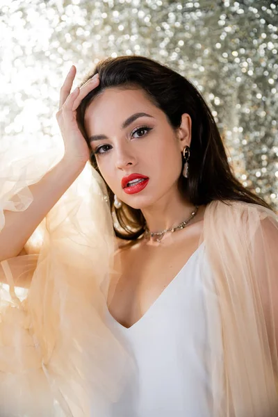 Brunette woman in elegant tulle dress holding hand near head and looking at camera on glitter background — Stock Photo