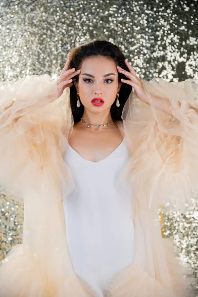 Mulher glamour em vestido elegante tocando cabelo morena e olhando para a câmera no fundo brilhante — Fotografia de Stock