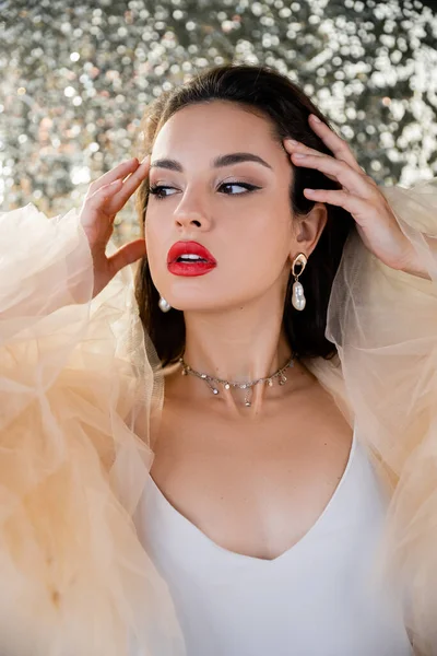 Elegant woman with red lips and pearl earrings looking away and touching hair on shiny background — Stock Photo