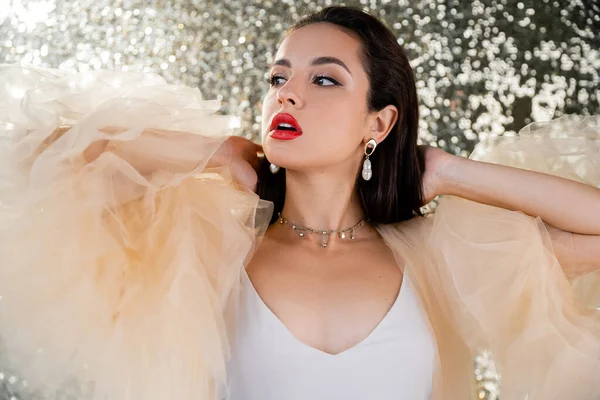 Mujer joven con labios rojos posando en elegante vestido de gasa y mirando hacia otro lado sobre un fondo brillante - foto de stock