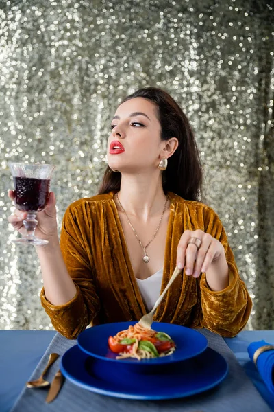 Mujer elegante con copa de vino tinto sosteniendo tenedor cerca de deliciosa pasta y mirando hacia otro lado sobre un fondo brillante - foto de stock