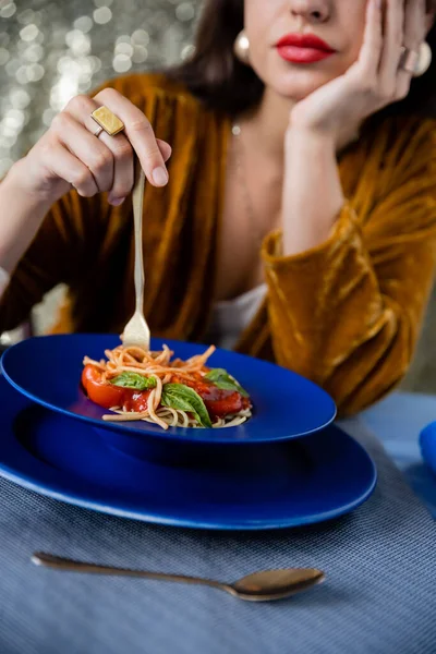 Vista recortada de la mujer borrosa sosteniendo tenedor cerca de placa azul con espaguetis sobre fondo brillante - foto de stock