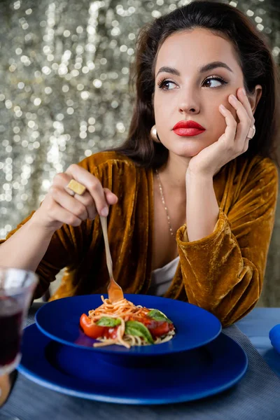 Mujer joven en vestido de terciopelo mirando hacia otro lado cerca de plato azul con espaguetis sobre fondo de brillo - foto de stock