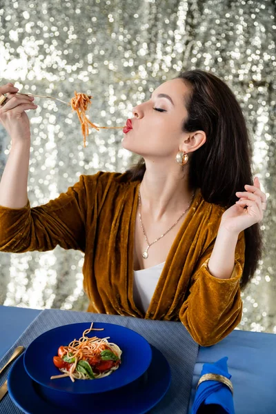 Brunette woman eating tasty spaghetti near blue plates on shiny silver background — Stock Photo