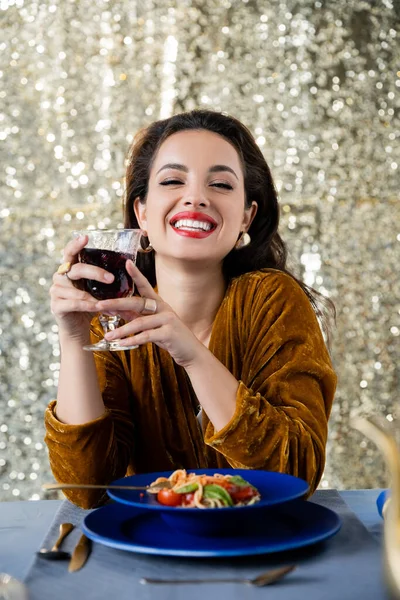 Mulher alegre e elegante com copo de vinho tinto olhando para a câmera perto de espaguete no fundo prata brilho — Fotografia de Stock