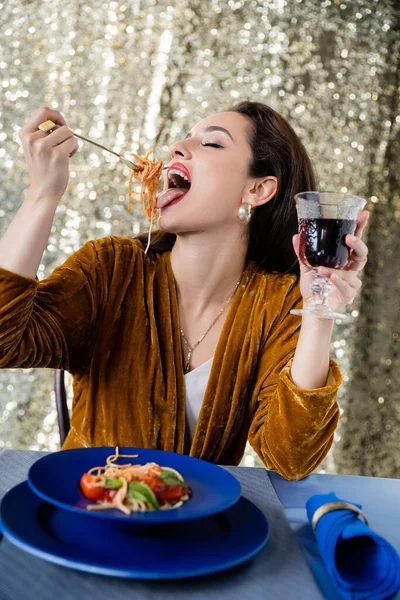 Jovem mulher em veludo vestido segurando vinho tinto e comer macarrão no fundo brilhante — Fotografia de Stock