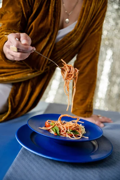 Vista ritagliata di donna in velluto vestito tenendo forchetta con pasta vicino piatti blu su sfondo lucido — Foto stock