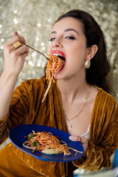 Mulher em veludo vestido comendo espaguete saboroso e olhando para a câmera no fundo de prata brilhante — Fotografia de Stock