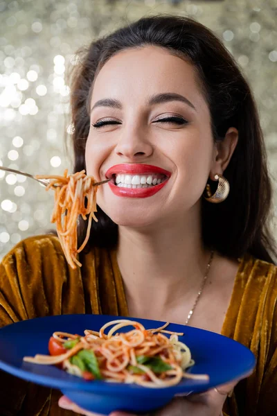 Fröhliche brünette Frau mit blauem Teller und Gabel mit Pasta auf glänzendem Hintergrund — Stockfoto