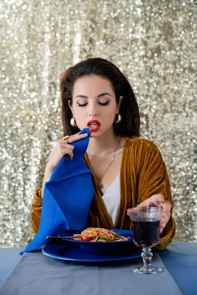 Femme sexy et élégante tenant serviette bleue près de délicieuses pâtes et verre de vin rouge sur fond de paillettes — Photo de stock