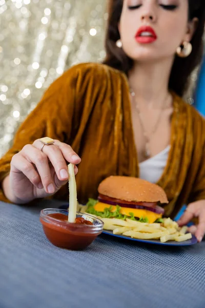 Mulher borrada em vestido de veludo mergulhando batatas fritas em tigela com ketchup perto de hambúrguer no fundo brilhante — Fotografia de Stock