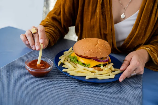 Vue recadrée d'une femme floue trempant des frites dans du ketchup près d'une assiette avec un délicieux hamburger sur fond gris — Photo de stock