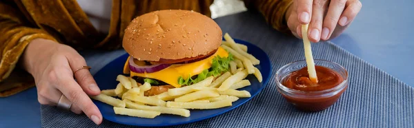 Partial view of woman dipping french fries into tomato sauce near delicious burger on blue plate, banner — Stock Photo