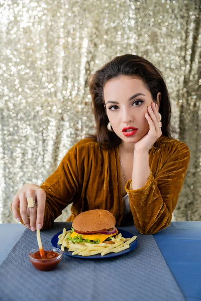 Femme brune en robe de velours tenant des frites près de ketchup et hamburger tout en regardant la caméra sur fond de paillettes — Photo de stock