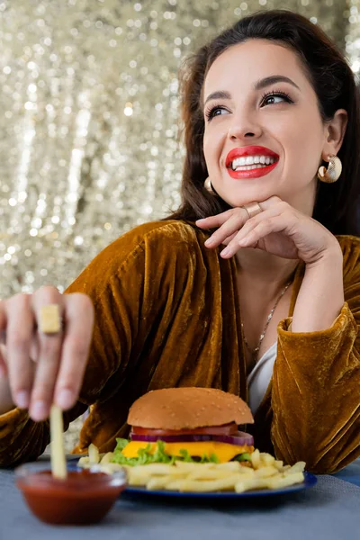 Mujer feliz y elegante sumergiendo papas fritas en ketchup cerca de deliciosa hamburguesa sobre un fondo brillante - foto de stock