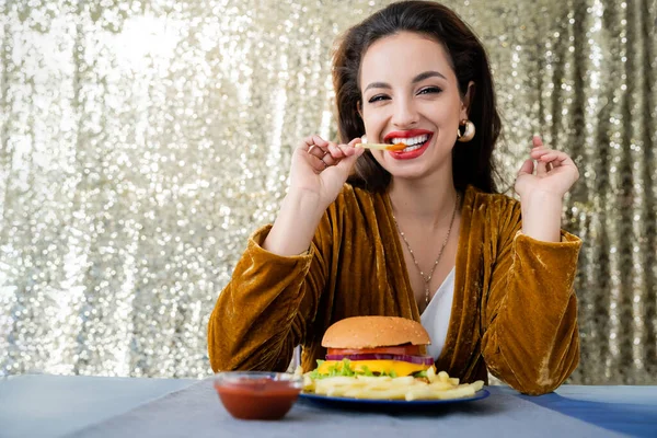 Стильная и веселая женщина смотрит в камеру и ест картошку фри рядом с бургером на блестящем серебряном фоне — стоковое фото