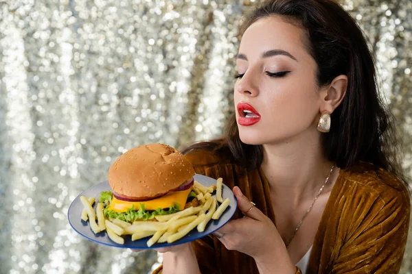 Mujer bonita y elegante sosteniendo plato azul con deliciosa hamburguesa y papas fritas sobre fondo brillante - foto de stock