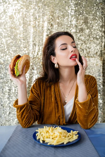 Seductora mujer con los ojos cerrados tocando el labio y sosteniendo hamburguesa cerca de papas fritas sobre fondo brillante - foto de stock