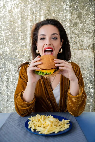 Брюнетка элегантная женщина смотрит в камеру во время еды гамбургер возле картошки фри на блестящем серебряном фоне — стоковое фото