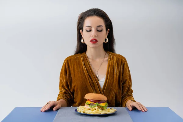 Femme élégante en robe de velours regardant assiette avec frites et hamburger sur table isolé sur gris — Photo de stock