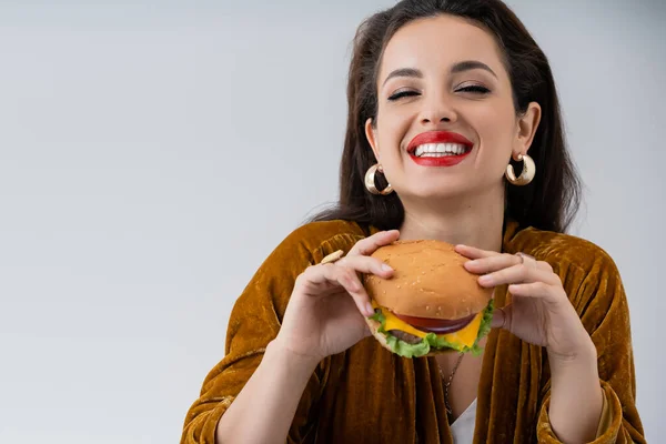 Mujer feliz en elegante vestido de terciopelo y pendientes de oro con deliciosa hamburguesa aislada en gris - foto de stock