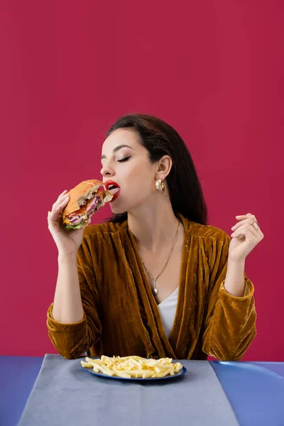 Femme sensuelle en robe de velours manger délicieux hamburger près des frites sur la table isolé sur rouge — Photo de stock