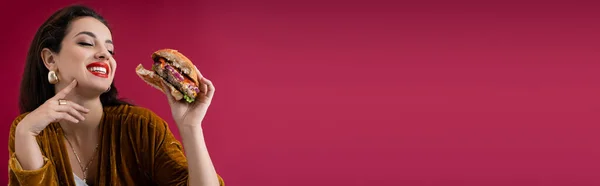 Joyful and fashionable woman holding tasty burger isolated on red, banner — Stock Photo