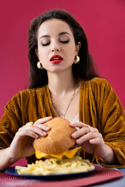 Sexy y elegante mujer mirando sabrosa hamburguesa cerca de papas fritas aisladas en rojo - foto de stock