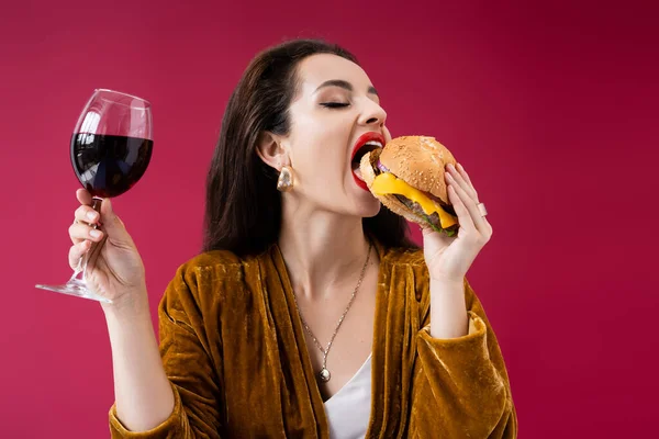 Brunette woman in velour dress holding glass of wine and eating tasty burger isolated on red — Stock Photo