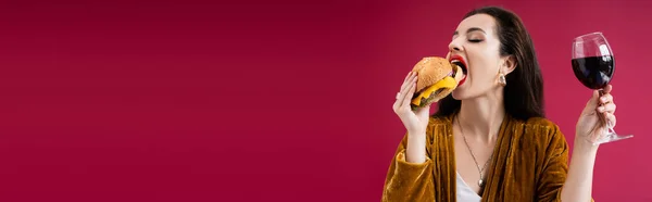 Morena mujer con copa de vino comiendo deliciosa hamburguesa aislada en rojo, pancarta - foto de stock