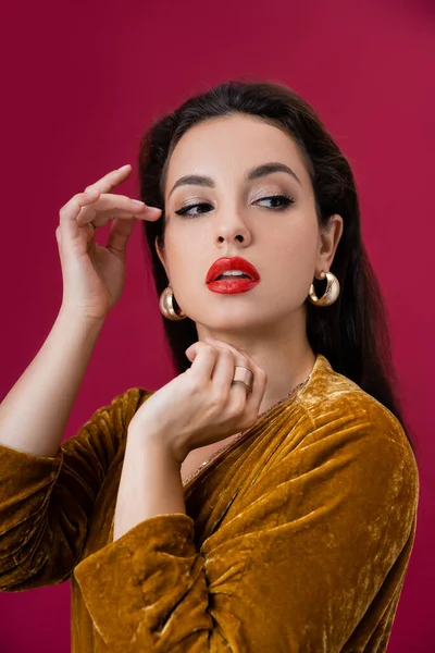 Mujer glamour en pendientes de oro y vestido de terciopelo agarrado de la mano cerca de la cara y mirando hacia otro lado aislado en rojo - foto de stock