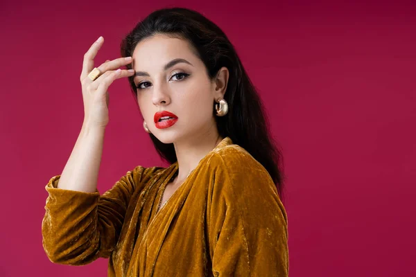 Charmante femme en robe de velours et boucles d'oreilles dorées touchant le visage et regardant la caméra isolée sur rouge — Photo de stock