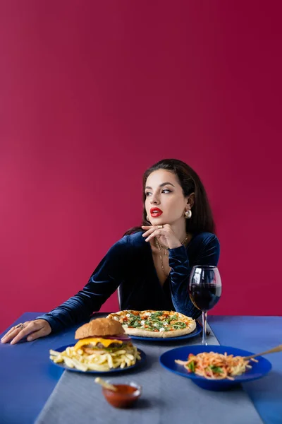 Femme brune en robe de velours regardant loin près de délicieux repas servi sur table bleue isolé sur rouge — Photo de stock